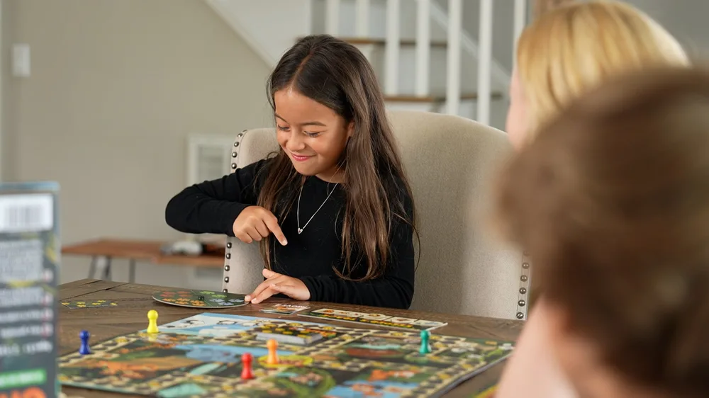 A young girl smiling while moving her pawn across the Keeper Chaos board at a family table