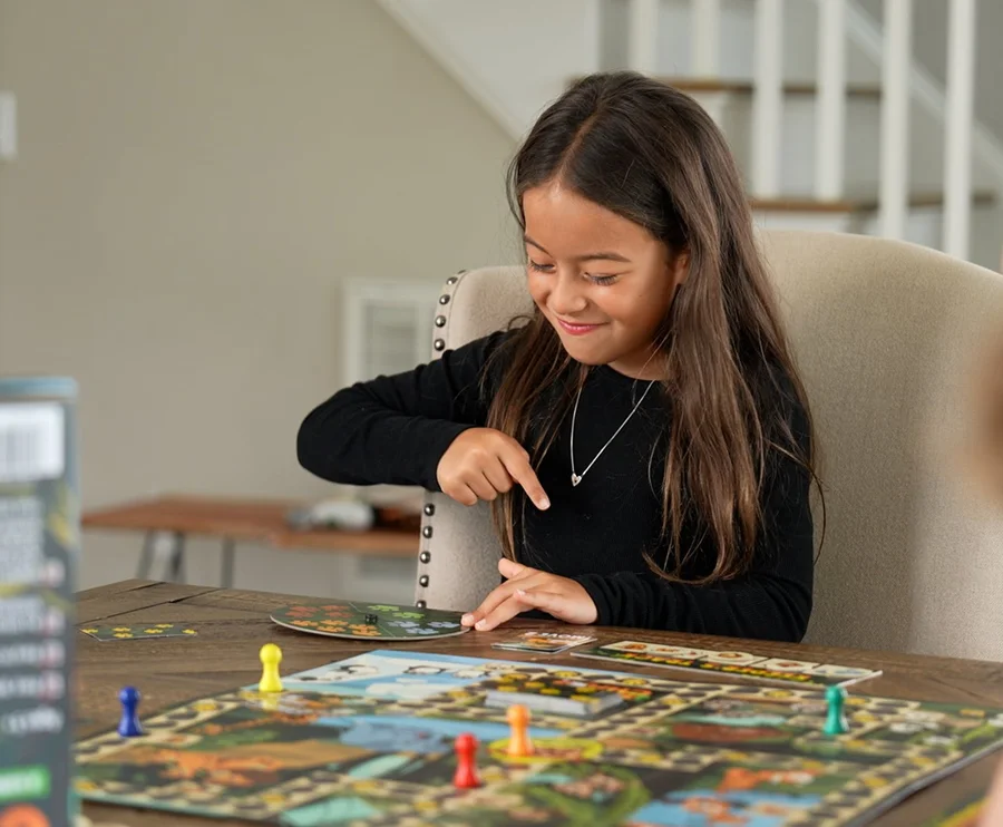 A girl smiling as she reaches for the spinner during a game of Keeper Chaos, the board and coloured pawns laid out on the table in front of her